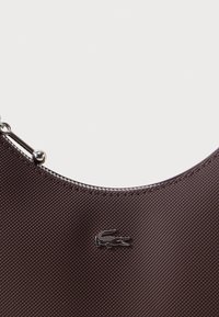 Hnědá nylónová taška s zakřiveným tvarem, vybavená stříbrným kováním a logem Lacoste. Viditelný texturovaný povrch a detail s uzávěrem na zip.
