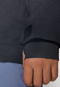 Black quilted fabric sleeve with textured finish, featuring a velcro patch and ribbed cuff, revealing a hand gripping the fabric.