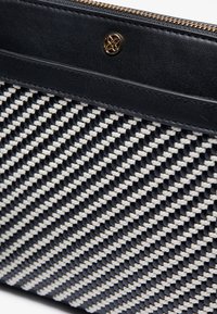 Svart vävd clutch med gräddvita detaljer, som har en texturerad textilväv och släta läderkanter, gulddragkedja och logotypdetalj.