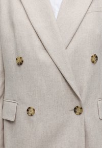 Blazer croisé beige avec quatre boutons en écaille et poches à rabat de chaque côté.