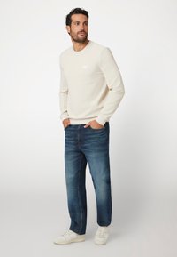Ljust beige sweatshirt med rundhals och litet logo, matchat med mörkblå avsmalnande jeans och vita sneakers. Slät tyg, avslappnad passform.