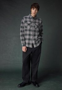 Cămașă din flanel gri și negru cu un model în carouri, având două buzunare pe piept, asortată cu pantaloni largi negri și cizme negre.