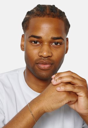 Ritratto di un uomo con capelli intrecciati che indossa una maglietta bianca. Mani giunte con un braccialetto d'oro sottile visibile al polso. Sfondo semplice.