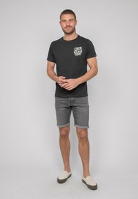 T-shirt noire à manches courtes avec un graphisme sur le devant, portée avec un short en denim gris retroussé. Le mannequin porte des chaussures légères à enfiler. Arrière-plan uni.