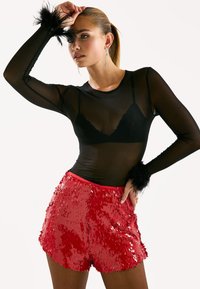 Haut noir transparent à manches longues avec des accents de plumes aux poignets, associé à un short rouge à paillettes scintillantes, créant un contraste audacieux de textures.