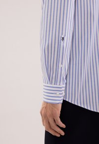 Niebiesko-biała koszula w paski z mankietem na guziki, z małym haftowanym kwiatowym detalem w pobliżu nadgarstka. Gładka faktura bawełnianej tkaniny.