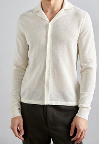 Cardigan bianco a maniche lunghe con un motivo a maglia testurizzata, chiusura frontale con bottoni e colletto classico. Indossato sopra pantaloni scuri e aderenti.