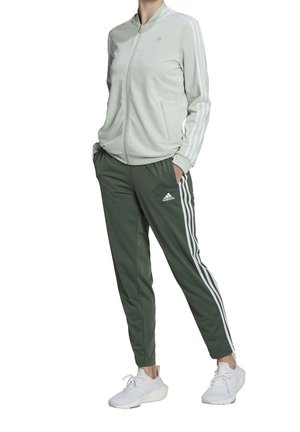 Personne portant une veste zippée gris clair et un pantalon de survêtement Adidas vert foncé avec des bandes blanches, associé à des baskets blanches.