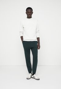 Sweatshirt gris clair avec des poignets côtelés, associé à un jogging vert foncé avec des chevilles élastiques, et des baskets blanches avec des accents noirs.
