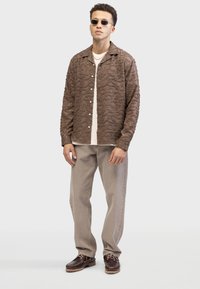 Brown textured shirt with wavy pattern, button closure, paired with a cream knit top, beige trousers, and brown shoes with laces.