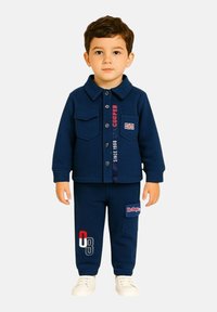 Ensemble pour enfant deux pièces bleu marine comprenant une chemise à boutons avec deux poches poitrine et un pantalon assorti avec des accents de logo, accompagné de baskets blanches.