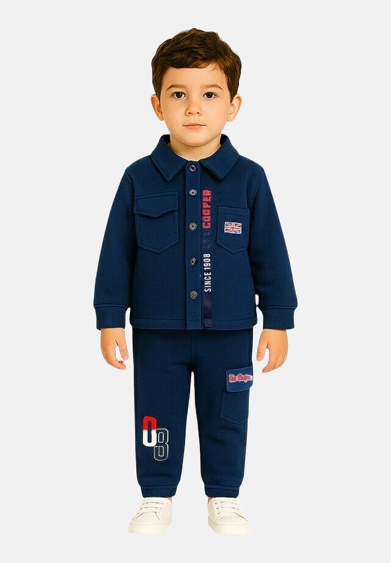 Ensemble pour enfant deux pièces bleu marine comprenant une chemise à boutons avec deux poches poitrine et un pantalon assorti avec des accents de logo, accompagné de baskets blanches.