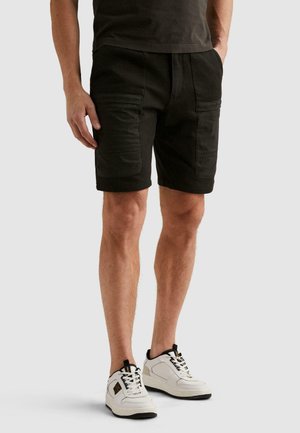 Mężczyzna ubrany w czarne krótkie spodenki typu cargo z wieloma kieszeniami, ciemną koszulę i białe sneakersy, stojący na jednolitym tle.