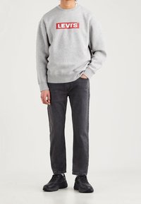 Sudadera gris con el logo rojo "LEVI'S", mezcla de algodón, sobre jeans negros. El modelo lleva zapatillas deportivas negras y está de pie contra un fondo liso.