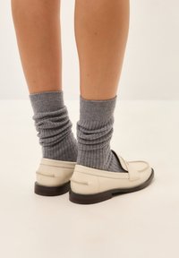 Loafers aus cremefarbenem Leder mit glatter Oberfläche und dunklen Gummisohlen, kombiniert mit gerippten grauen Wollsocken, die einen gestuften Look kreieren.