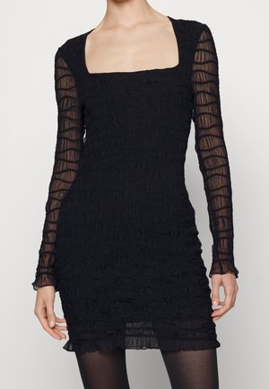 Schwarzes Mini-Kleid mit langen Ärmeln, geraffter Textur, transparenten Ärmeln, quadratischem Ausschnitt und gerafftem Saum, getragen von einer stehenden Person.