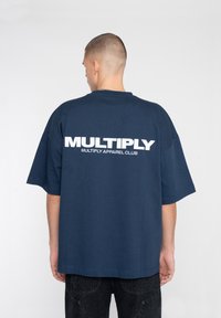 Vaatteena on merensininen ylisuuri t-paita, joka on valmistettu puuvillasta. T-paidassa on selässä rohkea valkoinen teksti "MULTIPLY", lyhyet hihat ja pyöreä kaula-aukko.