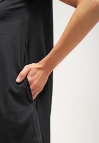 Svart klänning med avslappnad passform, med en sidoficka. Närbild av en hand som vilar på fickan, som visar den släta tygtexturen.