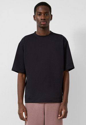 Jeune homme portant une chemise noire unie oversize à manches courtes et un pantalon rose clair sur un fond uni.