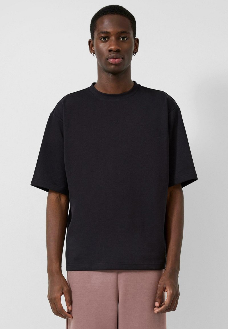 Jeune homme portant une chemise noire unie oversize à manches courtes et un pantalon rose clair sur un fond uni.