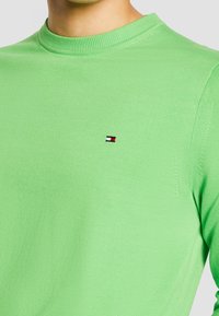 Personne portant un pull vert vif à col rond avec un petit logo rectangulaire rouge, blanc et noir sur la poitrine gauche.