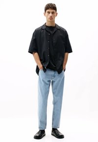 Cămașă neagră cu mânecă scurtă cu nasturi peste un tricou negru, asociată cu blugi albaștri deschis și pantofi negri slip-on. Design simplu, casual.