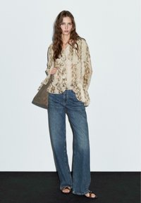 Beige ormönstrad blus med volangkant, tillsammans med högmidjade blå denimjeans. Modellen håller en grått tote-väska, framför en neutral bakgrund.