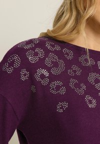 Lila tröja med silvriga nitar i leopardsmönster på axeln och övre bröstet, bärs av en person med blont hår.