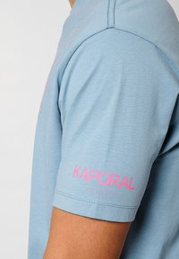 Lichtblauwe T-shirt met korte mouwen van katoen; heeft een roze "KAPORAL" logo op de linkermouw; zachte textuur, casual pasvorm.