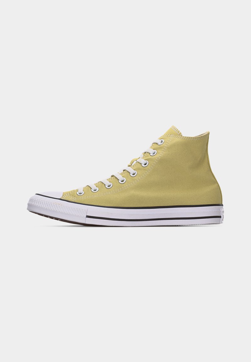 Zapatilla de lona beige de caña alta con suela y puntera de goma blanca, cordones blancos y detalle de raya negra a lo largo de la suela.