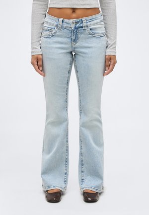 Žena stojaca pred bielym pozadím, oblečená v svetlomodrých zvonových rifliach, sivom dlhom rukáve s odhaleným bruškom a tmavohnedých topánkach so štvorcovou špičkou.