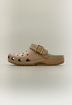 Μπεζ κλογκ τύπου slip-on με οπές αερισμού και ρυθμιζόμενο λουράκι με χρυσή πόρπη σε απλό ουδέτερο φόντο.