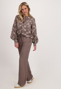 Vrouw in een losse bruine blouse met bloemenprints en bijpassende wijde broek, met witte sneakers, staand tegen een effen achtergrond.