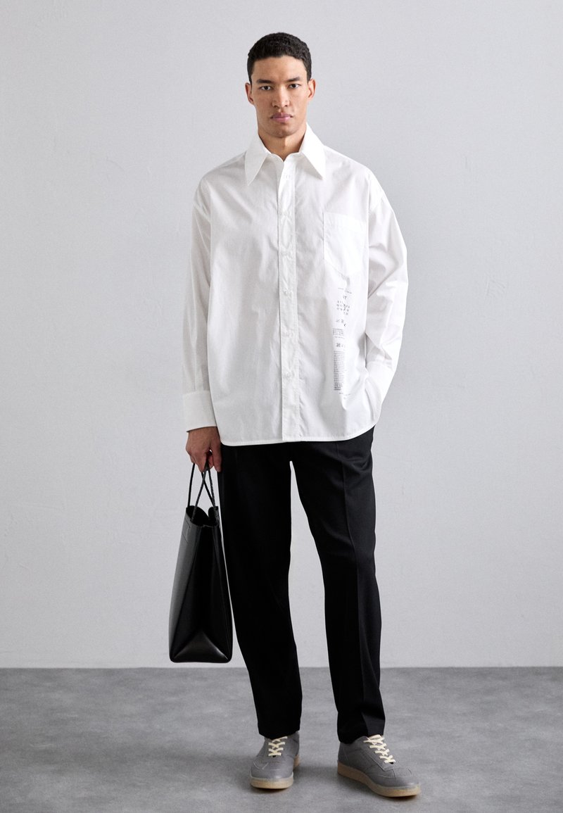 Hvid oversized skjorte med knapper og en brystlomme, parret med sorte bukser og grå sneakers. Modellen holder en sort tote bag.