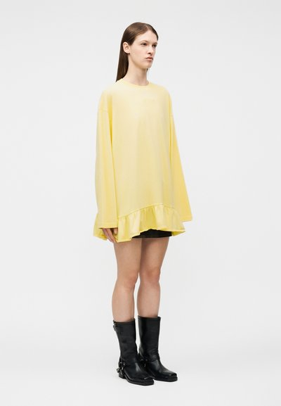 Νεαρή γυναίκα στέκεται φορώντας μια υπερμεγέθη μακρυμάνικη μπλούζα σε ανοιχτό κίτρινο με βολάν στο τελείωμα, μαύρο σορτς και μαύρες μπότες μέχρι τη μέση της γάμπας σε λευκό φόντο.