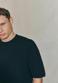 Młody mężczyzna z krótkimi, falującymi włosami, ubrany w czarną koszulkę, spogląda nieco na bok na neutralnym tle.