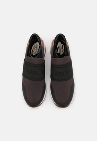 Chaussures de sport noires et marron avec une tige textile foncée, une sangle élastique ornée d'un logo et une semelle en caoutchouc avec accents texturés.