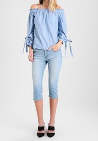 Carmenbluse mit Off-Shoulder, blauen Streifen und grünen Akzenten, lange Ärmel, die an den Handgelenken gebunden sind; kombiniert mit hellblauen, knöchellangen Jeans und schwarzen High Heels.