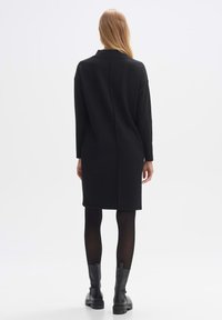 Schwarzes, midilanges Kleid mit lockerer Passform, langen Ärmeln und einer Naht im Rücken. Trägt es über schwarzen Strumpfhosen und klobigen schwarzen Stiefeln. Glatte Textur.