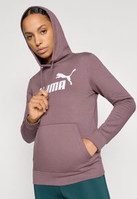 Mikina s dlhým rukávom a kapucňou vo vyblednutej fialovej farbe s bielym logom Puma. Obsahuje predné vrecko a rebrované manžety pre priliehavý vzhľad.