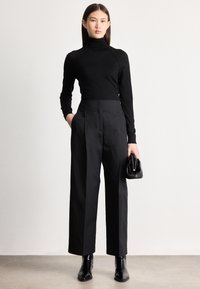 Γυναίκα στέκεται φορώντας μαύρο ζιβάγκο, μαύρο παντελόνι ψηλόμεσο με φαρδιά γραμμή, μαύρα μποτάκια μέχρι τον αστράγαλο, κρατώντας μια μικρή μαύρη τσάντα, μπροστά σε απλό λευκό φόντο.