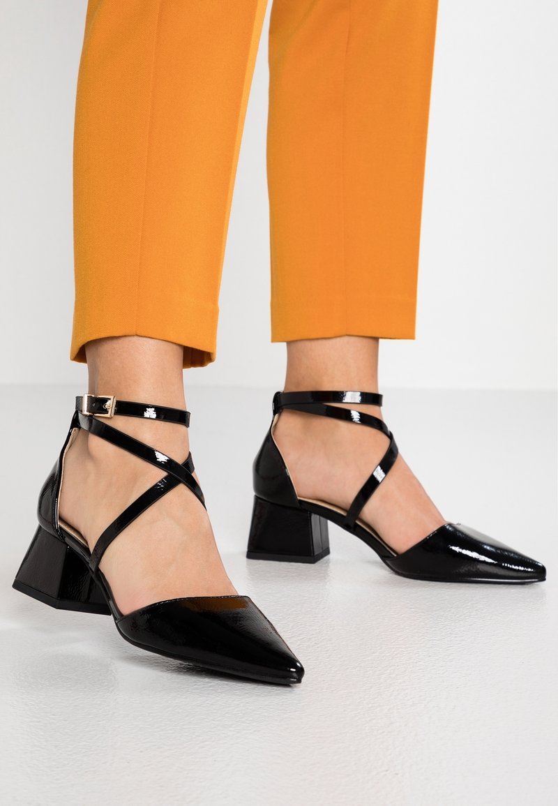 Escarpins à bouts pointus en cuir verni noir avec brides croisées à la cheville, portés avec un pantalon orange raccourci sur un sol blanc lisse.