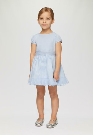 lichtblauwe katoenen jurk met korte pofmouwen, gathered taille, kantafwerking aan de zoom, en zilveren platte schoenen met bloemenaccenten.