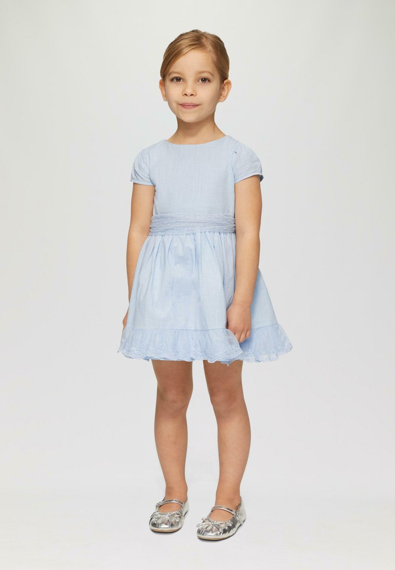 Helles blaues Baumwollkleid mit kurzen Puffärmeln, gerafftem Tailleneinsatz, Spitze am Saum und silbernen flachen Schuhen mit floralem Akzent.