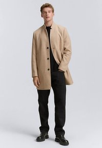 Bež plašč iz volnenega mešanega materiala s stoječim ovratnikom, petimi gumbi in stranskimi žepi, nošen čez črno srajco in temne hlače, kombiniran s črnimi škornji.