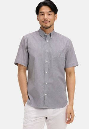 Homme portant une chemise à rayures noires et blanches à manches courtes avec col, debout les mains dans les poches d'un pantalon blanc.