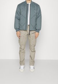 Teal quiltad bomberjacka med ribbade ärmar, tillsammans med bruna cargobukser med flera fickor och vita sneakers.