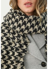 Szalik w wzór houndstooth w czerni i kremie, wykonany z miękkiej tkaniny, z frędzlami na brzegach, zarzucony na szary płaszcz.