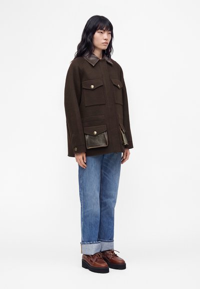 Personne portant une veste marron foncé avec des poches et un col en cuir, un jean bleu avec revers et des chaussures à plateforme marron, debout sur un fond uni.