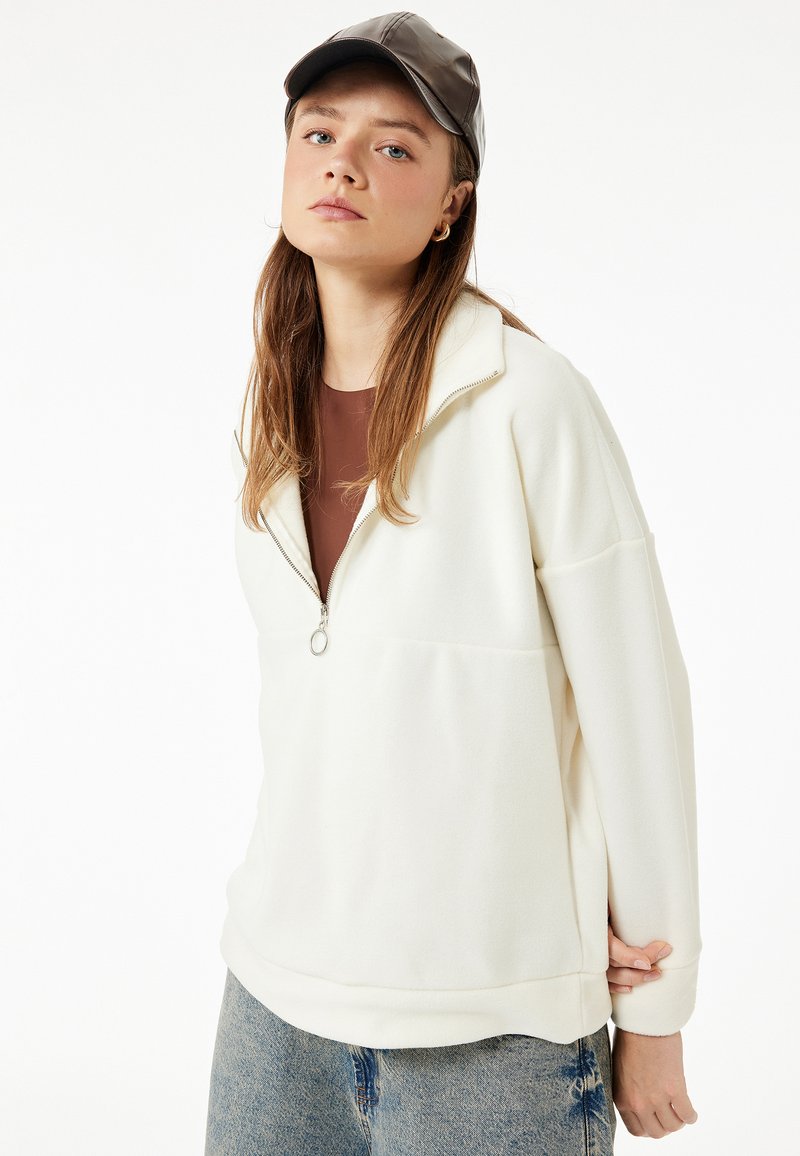 Sudadera de felpa blanca con cuello de cremallera de cuarto, diseño de hombros caídos, tejido texturizado y un ajuste relajado. Combinada con jeans de tela vaquera azul.